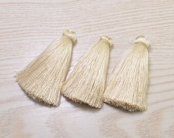 May include: Three cream-colored silk tassels. Each tassel has a rounded top and a long, fringed bottom. The tassels are arranged in a row, with the light-colored background providing a neutral backdrop.