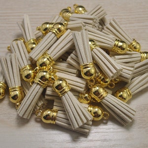 Könnte beinhalten: Ein Haufen beigefarbener Wildlederquasten mit goldfarbenen Metallkappen und Schlaufen. Die Quasten sind zusammengelegt, wobei die goldenen Kappen das Licht reflektieren. Geeignet für Schmuckherstellung oder Bastelprojekte.