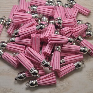 Puede incluir: Un montón de borlas de gamuza rosa con tapas de metal plateado. Las borlas son todas del mismo tamaño y forma, y están dispuestas en un patrón aleatorio.
