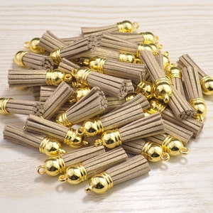 Könnte beinhalten: Ein Haufen beigefarbener Wildlederquasten mit goldfarbenen Metallkappen und Schlaufen. Die Quasten sind zusammengefasst und zeigen ihr strukturiertes, gefranstes Design. Die goldenen Kappen verleihen dem Bastelbedarf einen Hauch von Eleganz.