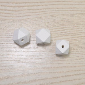 Może przedstawiać: Trzy białe geometryczne drewniane koraliki o fasetowym wzorze. Każdy koralik ma otwór wywiercony pośrodku. Koraliki ułożone są na jasnej powierzchni z usłojeniem drewna.