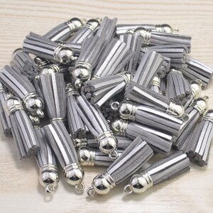 Könnte beinhalten: Ein Haufen silbergrauer Wildleder-Quasten mit silbernen Metallkappen und Schlaufen. Die Quasten sind etwa 5 cm lang.