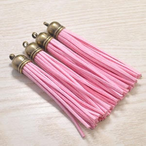 Könnte beinhalten: Vier rosa Wildlederquasten mit antik-bronze farbenen Kappen. Die Quasten sind diagonal angeordnet und zeigen ihre weiche Textur und lebendige Farbe. Ideal für Schmuckherstellung oder Bastelprojekte.