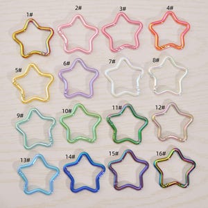 May include: A collection of sixteen star-shaped metal keychains in various colors, including gold, pink, purple, green, and blue. Each keychain has a clasp for attaching to keys or other items. The keychains are numbered 1 through 16.