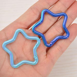 May include: Two star-shaped carabiner clips in iridescent blue and blue hues. The clips are held in a hand, showcasing their metallic finish and the spring-loaded gate for easy attachment. These are ideal for keychains or crafting.
