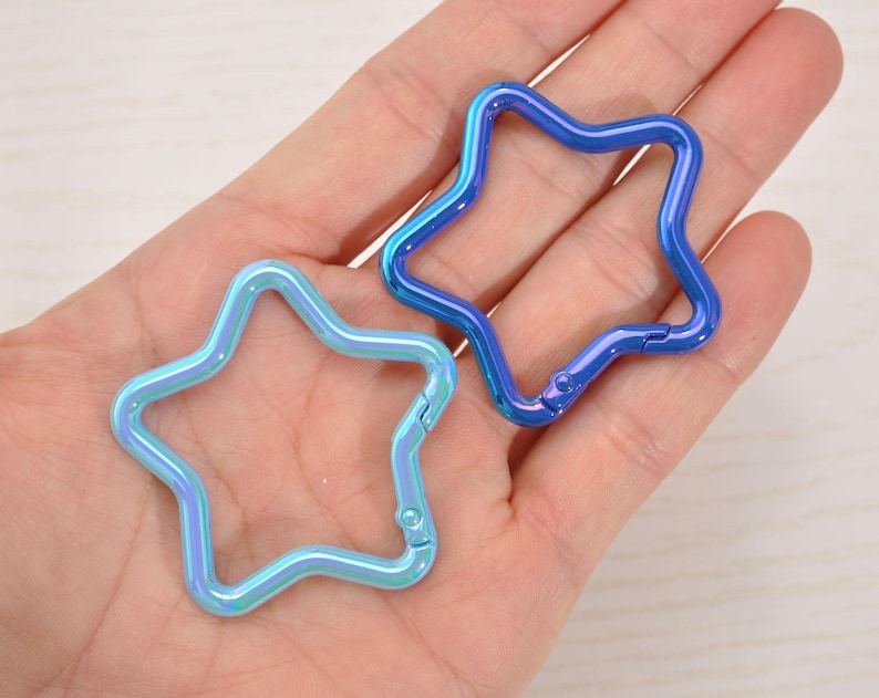 May include: Two star-shaped carabiner clips in iridescent blue and blue hues. The clips are held in a hand, showcasing their metallic finish and the spring-loaded gate for easy attachment. These are ideal for keychains or crafting.