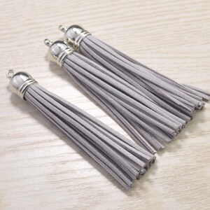 Könnte beinhalten: Drei graue Wildleder-Quasten mit silbernen Metallkappen. Die Quasten sind etwa 12,5 cm lang.