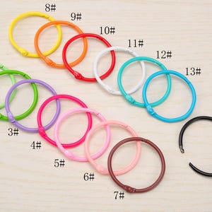 Könnte beinhalten: Eine Sammlung von vierzehn losen Metall-Buchringen in verschiedenen Farben, darunter Gelb, Orange, Rot, Weiß, Türkis, Blau, Hellgrün, Grün, Lila, Magenta, Pink, Hellrosa, Braun und Schwarz, jeweils beschriftet mit einer Nummer von 1# bis 14#.