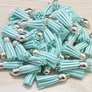 Puede incluir: Un montón de borlas de gamuza azul claro con tapas de metal plateado. Las borlas miden aproximadamente 5 cm de largo.