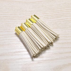 Puede incluir: Un conjunto de cinco borlas de color crema con tapas de metal dorado. Las borlas están hechas de fibras suaves y esponjosas.
