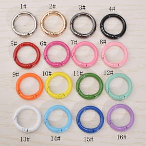 Puede incluir: Una colección de dieciséis anillos redondos de metal con resorte en varios colores, incluyendo plata, oro, negro, rojo, rosa, naranja, amarillo, verde, azul claro, azul y morado. Cada anillo tiene un cierre de resorte.