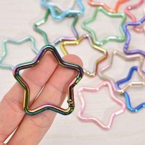 May include: A hand holds a star-shaped iridescent metal clasp. Other star-shaped clasps in various colors, including pink, blue, green, and orange, are scattered in the background. The clasp has a spring-loaded gate for easy attachment.