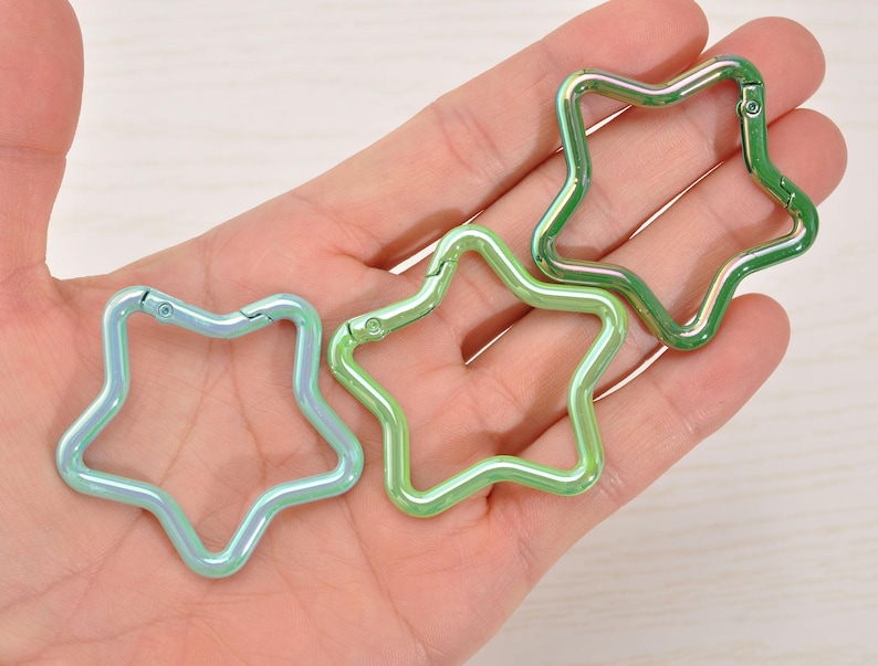 May include: Three star-shaped carabiner clips in iridescent green and blue hues. The clips are made of metal and have a spring-loaded gate for easy attachment. These decorative clips can be used for keychains, bag charms, or other craft projects.