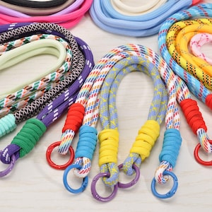 May include: A collection of colorful rope lanyards with various patterns and colors, including pink, blue, yellow, and purple. Each lanyard has a metal ring and a contrasting color grip.