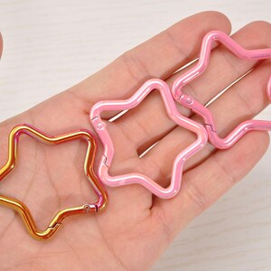 May include: Three star-shaped carabiner clips in gold and pink. The gold clip has a gradient effect, transitioning from gold to pink. The pink clips have a pearlescent finish. All clips have a hinged design for easy attachment.