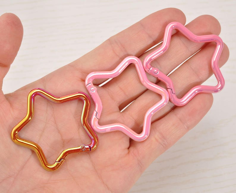 May include: Three star-shaped carabiner clips in gold and pink. The gold clip has a gradient effect, transitioning from gold to pink. The pink clips have a pearlescent finish. All clips have a hinged design for easy attachment.