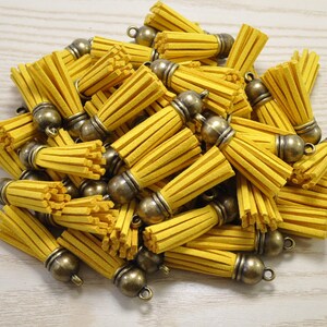 Puede incluir: Un montón de borlas de gamuza amarilla con tapas y lazos de color bronce antiguo. Las borlas son cilíndricas con ranuras verticales, y las tapas tienen forma redondeada. Estas borlas decorativas son adecuadas para la fabricación de joyas y proyectos de artesanía.