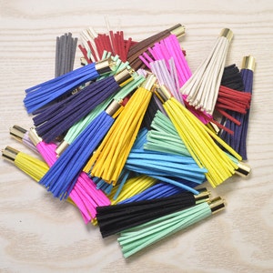 Könnte beinhalten: Eine Sammlung bunter Quasten in verschiedenen Farbtönen, darunter Blau, Gelb, Rosa und Grün. Jede Quaste hat eine goldfarbene Metallkappe. Die Quasten bestehen aus einem weichen, gefransten Material.