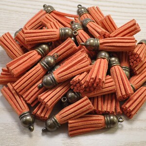 Puede incluir: Un montón de borlas de ante naranja con tapas y lazos de bronce antiguo. Las borlas están agrupadas, mostrando su diseño texturizado y con flecos. Estas borlas decorativas son adecuadas para la fabricación de joyas, la artesanía o la decoración.