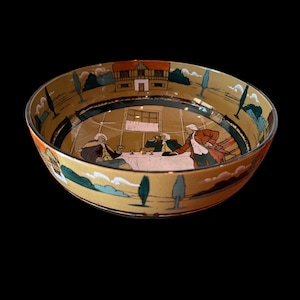 May include: A decorative bowl with a yellow interior and a black exterior. The bowl features a painted scene of people at a table, a building, and trees. The rim of the bowl is red. The bowl is likely vintage.
