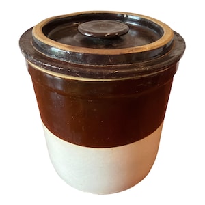 May include: A cylindrical stoneware crock featuring a two-tone design with a dark brown glazed upper section and a light cream-colored base. The container includes a matching dark brown lid with a central knob handle and a smooth, glossy finish.
