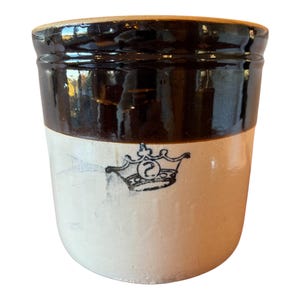 May include: A cylindrical stoneware crock featuring a two-tone design with a dark brown glazed upper section and a light beige lower section. A black stamped graphic of a crown with the letter S inside is centered on the beige body.