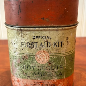 May include: Vintage metal first aid kit with a red lid and a cylindrical body. The body is painted with green, orange, and cream colors, and features the text "OFFICIAL FIRST AID KIT" and "BOY SCOUTS OF AMERICA".