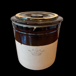 May include: A vintage stoneware crock with a dark brown glaze and a cream-colored base. The crock features a lid and a decorative crown emblem with the number 2. The antique kitchenware is set against a black background.