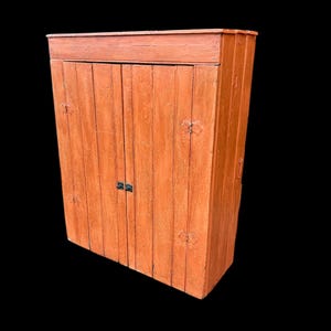 May include: A tall, orange-painted wooden cabinet with two doors and decorative metal hardware. The cabinet has a rectangular shape and a simple design, suitable for storage. The doors are closed, and the cabinet is set against a black background.