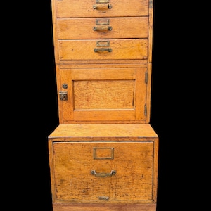 May include: A tall, light brown wooden filing cabinet featuring three small upper drawers with brass label holders and handles, a central cabinet door with a latch, and one large bottom drawer with a brass handle, all supported by four light-colored legs.