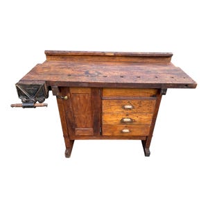 May include: A weathered brown wooden workbench featuring a large rectangular top with multiple square holes, a black metal side vise with a wooden handle, a single cabinet door on the left, and three stacked drawers on the right with metal pulls.