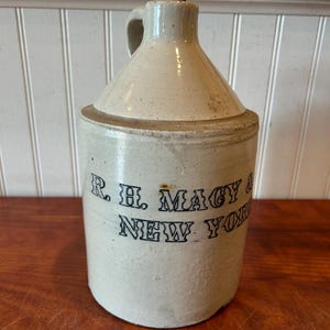May include: A vintage stoneware jug with a handle and a cork stopper. The jug is off-white with a dark blue inscription that reads "R. H. Macy & New York". The jug is cylindrical with a rounded top.