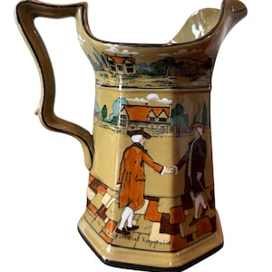 May include: A vintage ceramic pitcher with a tan background and a detailed illustration. The pitcher features a handle and a spout. The illustration depicts two figures shaking hands, with a landscape scene of houses and trees. The base has a geometric pattern.