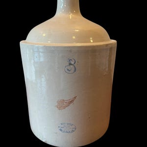 May include: Off-white stoneware jug with a rounded top and a narrow spout. The jug features a blue number "3", a brown wing emblem, and a blue stamp with text. The jug is likely an antique or vintage piece.