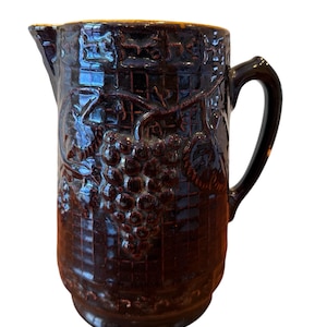May include: A dark brown ceramic pitcher with a handle and spout. The pitcher features a raised relief design of grapes and leaves. The surface has a textured, grid-like pattern. The interior rim has a golden-brown color.