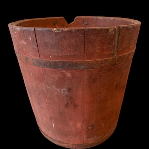 May include: A weathered, red wooden bucket with a slightly oval top and a metal band around the middle. The bucket has a rustic appearance, with visible wood grain and signs of age. The background is black.
