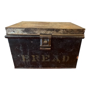 May include: A rectangular metal bread box with a dark brown finish and a weathered, metallic lid. The front of the container features the word BREAD in large, block lettering. A central metal latch secures the lid to the main body.