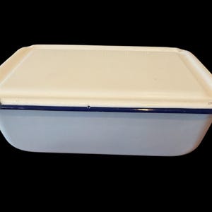 May include: A rectangular white enamelware container with a lid. The container has a blue trim and shows signs of wear around the edges. The lid is slightly off-white. The container is likely used for food storage.