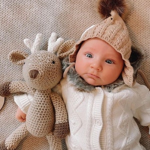May include: A baby in a beige knitted hat with a faux fur trim and a white cable knit outfit. A crocheted reindeer toy with brown and white details lies next to the baby on a textured surface.