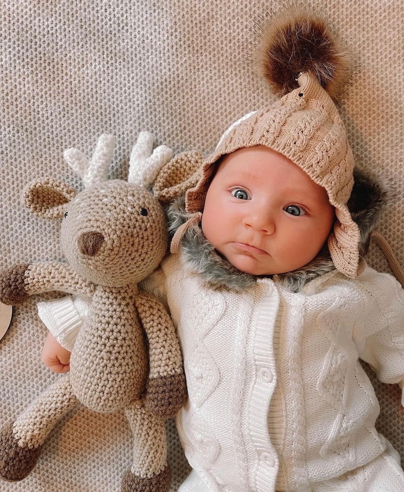 May include: A baby in a beige knitted hat with a faux fur trim and a white cable knit outfit. A crocheted reindeer toy with brown and white details lies next to the baby on a textured surface.