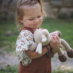May include: A young child in a brown overall and floral top, holding a beige and brown crocheted toy. The child has blonde hair and a brown bow. The background is a blurred green.