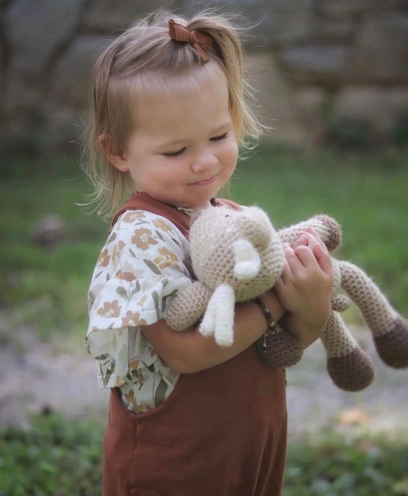May include: A young child in a brown overall and floral top, holding a beige and brown crocheted toy. The child has blonde hair and a brown bow. The background is a blurred green.