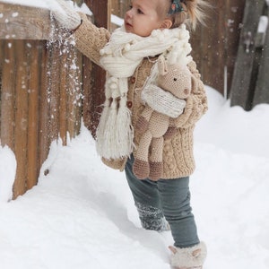 May include: A child in a beige cable-knit cardigan, white scarf, and mittens plays in the snow, holding a crocheted horse toy. The scene is set outdoors with a wooden fence and falling snow. The child is wearing grey trousers.