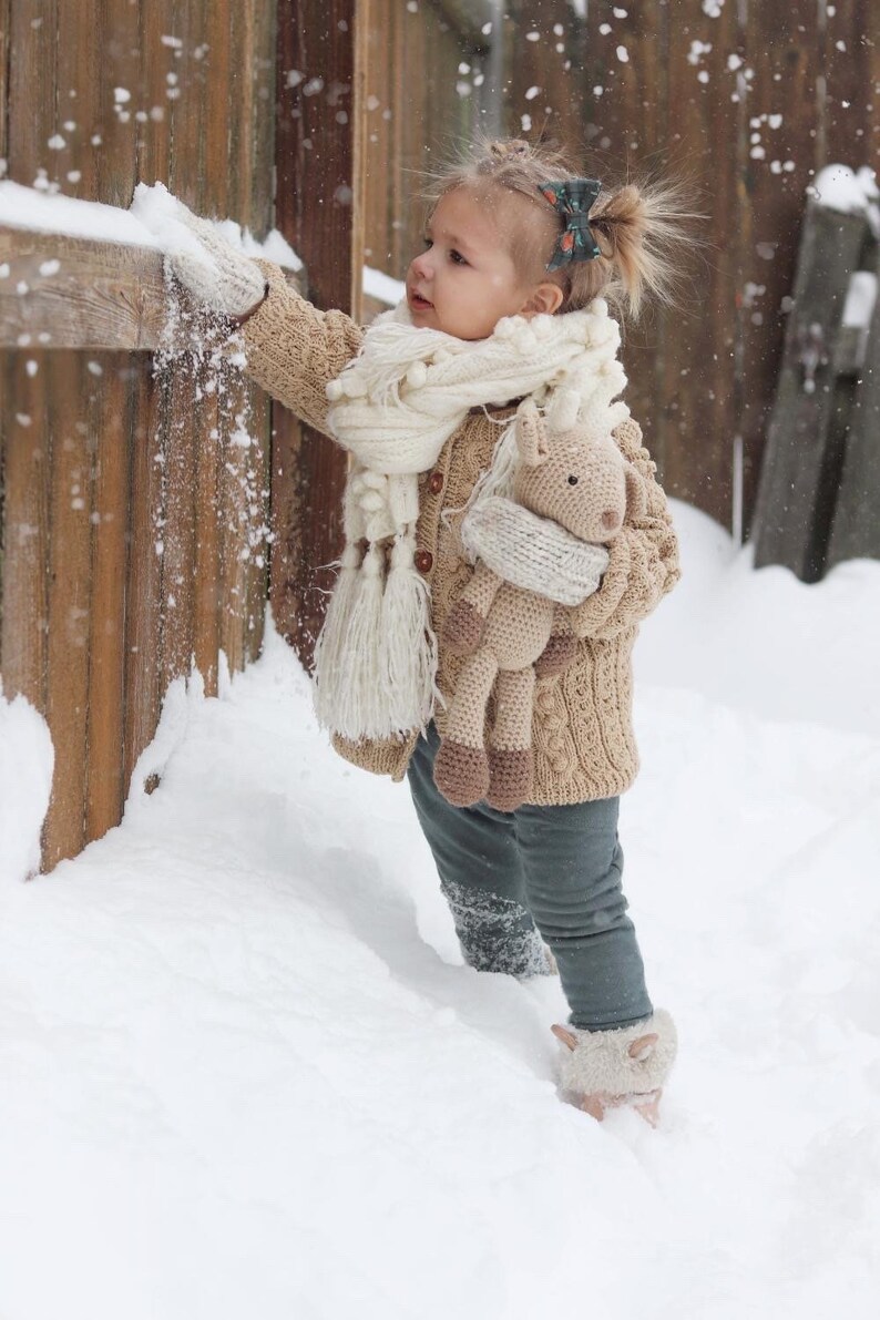 May include: A child in a beige cable-knit cardigan, white scarf, and mittens plays in the snow, holding a crocheted horse toy. The scene is set outdoors with a wooden fence and falling snow. The child is wearing grey trousers.