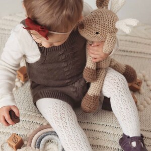 May include: A child in a brown romper, white tights, and purple shoes holds a crocheted reindeer toy. The romper features a cable knit pattern. Wooden blocks and a rainbow decoration are also visible.