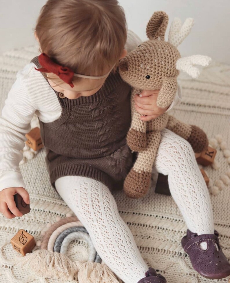 May include: A child in a brown romper, white tights, and purple shoes holds a crocheted reindeer toy. The romper features a cable knit pattern. Wooden blocks and a rainbow decoration are also visible.