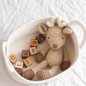 May include: A crocheted deer toy with brown details rests in a white woven basket with a handle. Wooden blocks spelling "L I V E R" are scattered around the deer. The deer has cream-coloured antlers and brown hooves.