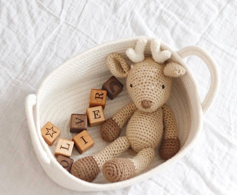 May include: A crocheted deer toy with brown details rests in a white woven basket with a handle. Wooden blocks spelling "L I V E R" are scattered around the deer. The deer has cream-coloured antlers and brown hooves.