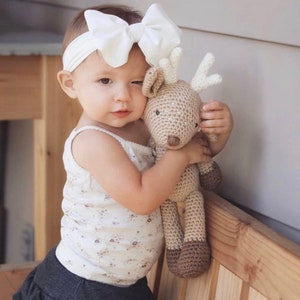 May include: A young child wearing a white bow headband and a floral print tank top, holding a handmade crochet deer plush toy. The deer is beige with brown accents and white antlers. The child is wearing a denim skirt.