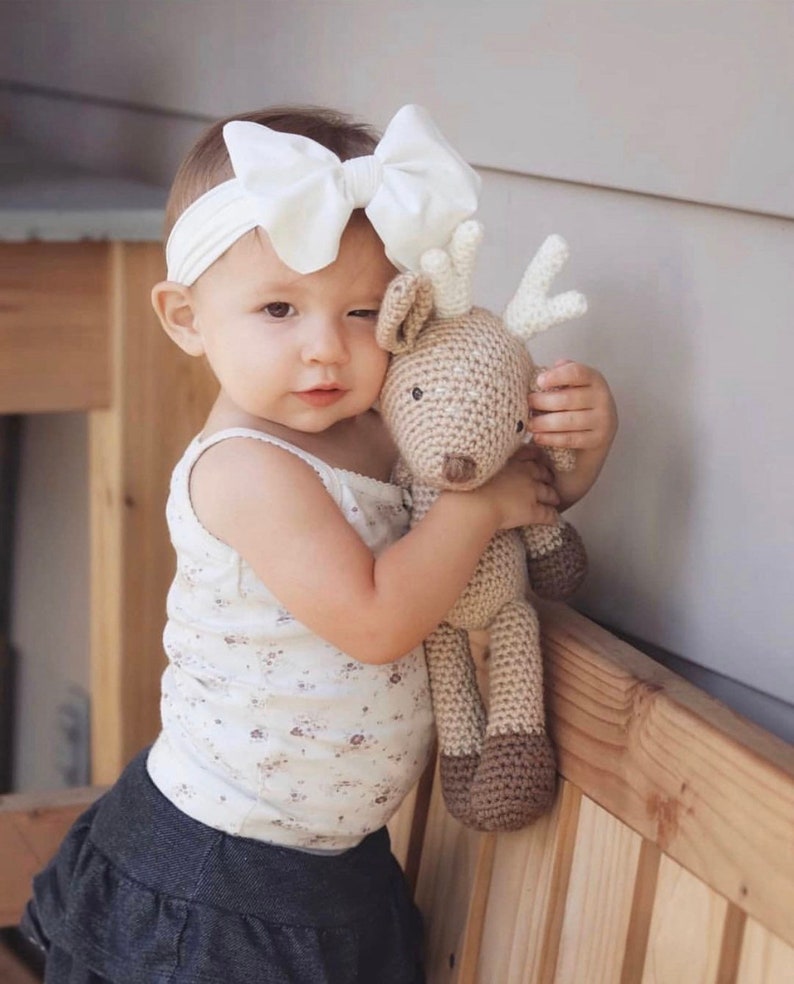 May include: A young child wearing a white bow headband and a floral print tank top, holding a handmade crochet deer plush toy. The deer is beige with brown accents and white antlers. The child is wearing a denim skirt.